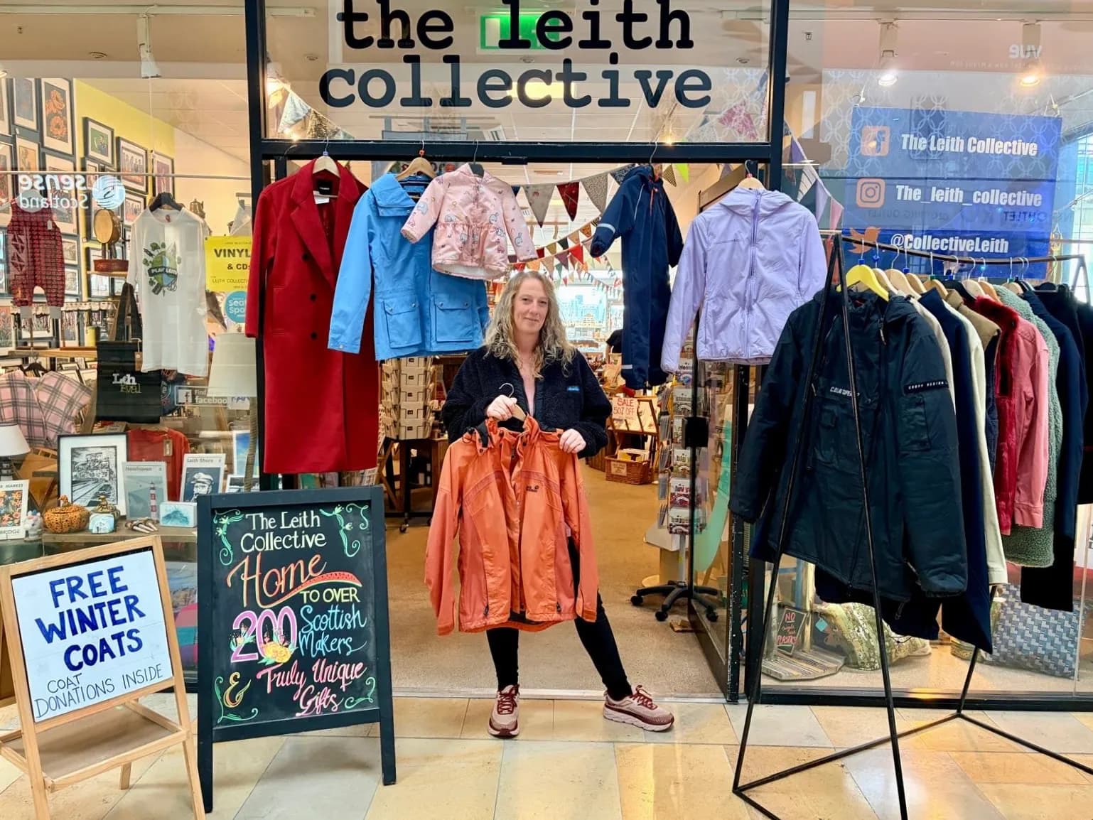 A woman stands in the doorway of "The Leith Collective" shop, holding up two orange jackets, surrounded by racks of clothing and signs for free winter coats and Scottish makers.