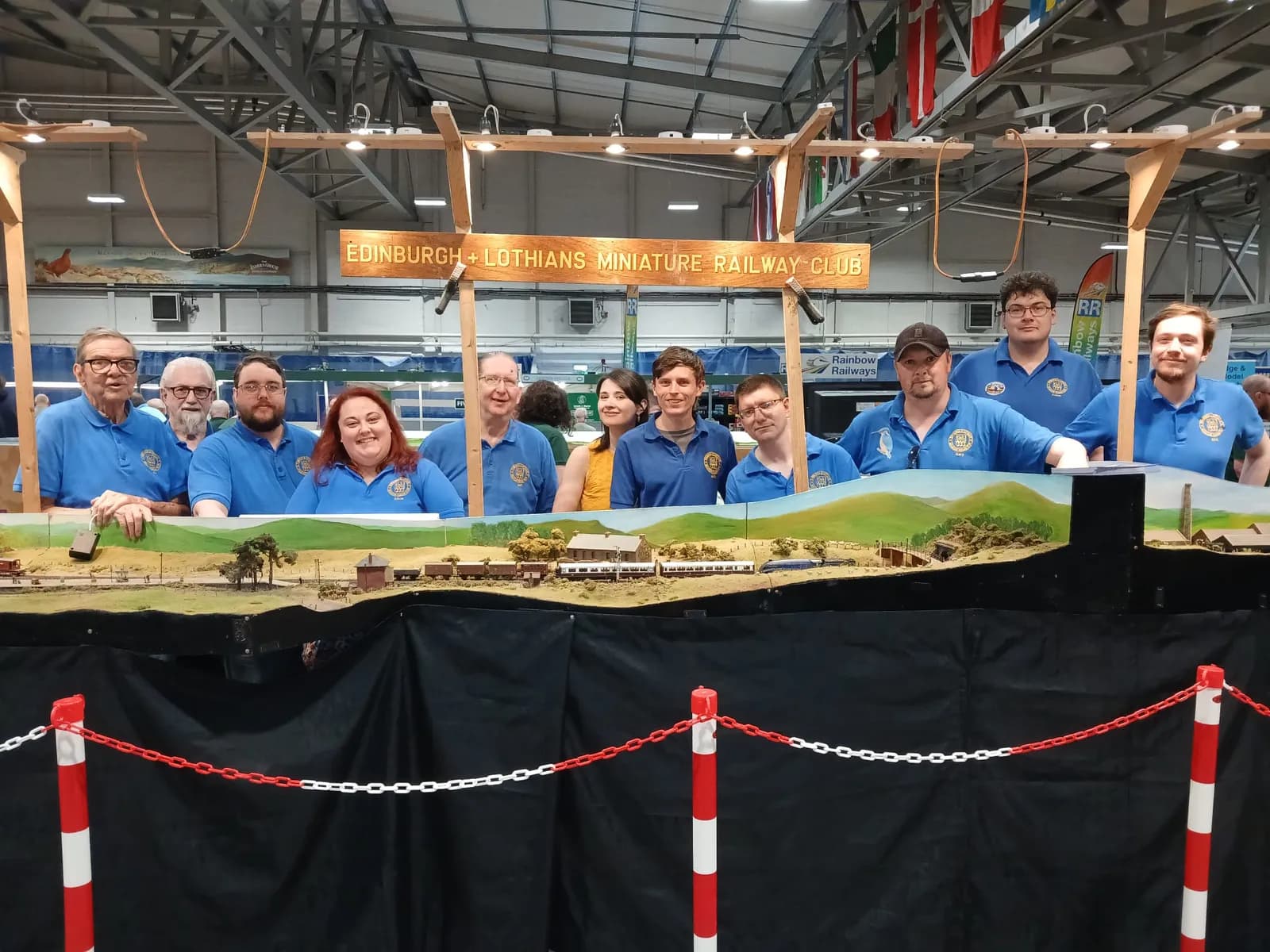 A group of people in blue shirts are gathered behind a detailed miniature train set, with a sign above them reading "Edinburgh & Lothians Miniature Railway Club."