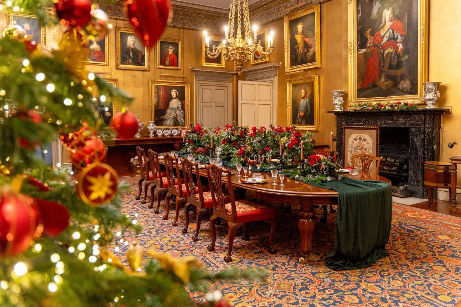 A long dining table, richly decorated with red flowers and greenery, is set for Christmas dinner in a grand hall with portraits on the walls.