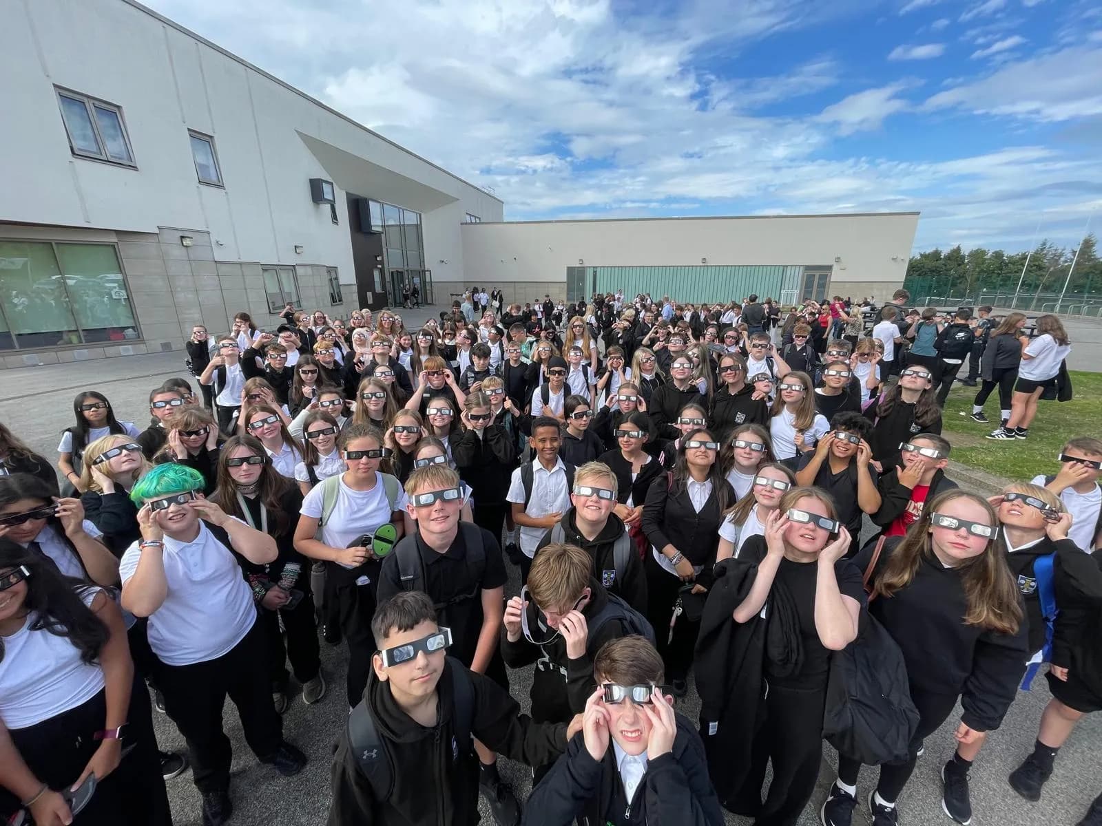 A large group of students wearing protective glasses and looking upwards, standing outside a modern school building under a cloudy sky.