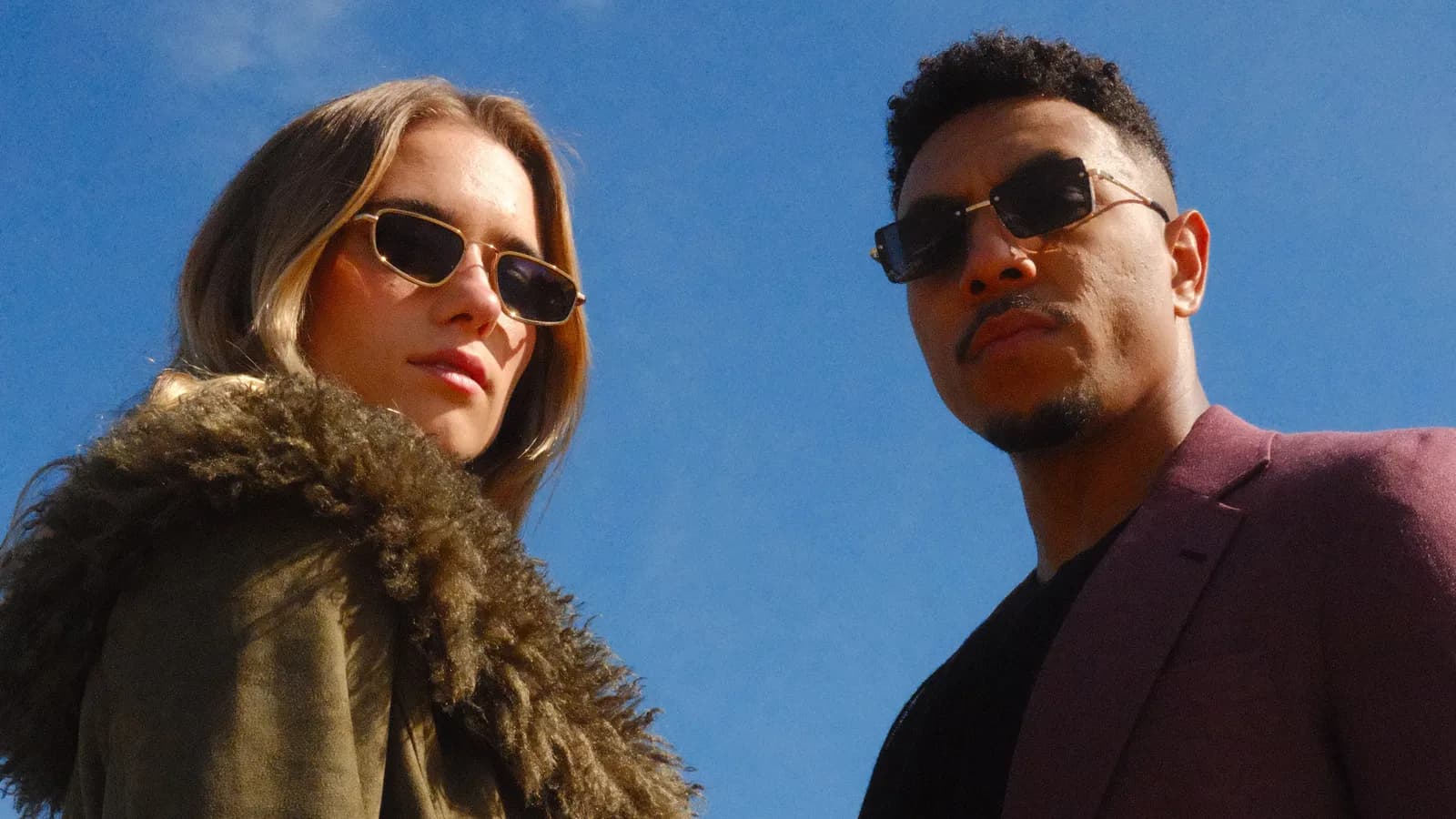 A woman in a green fur-trimmed jacket and a man in a maroon suit, both wearing sunglasses, pose outdoors against a clear blue sky.