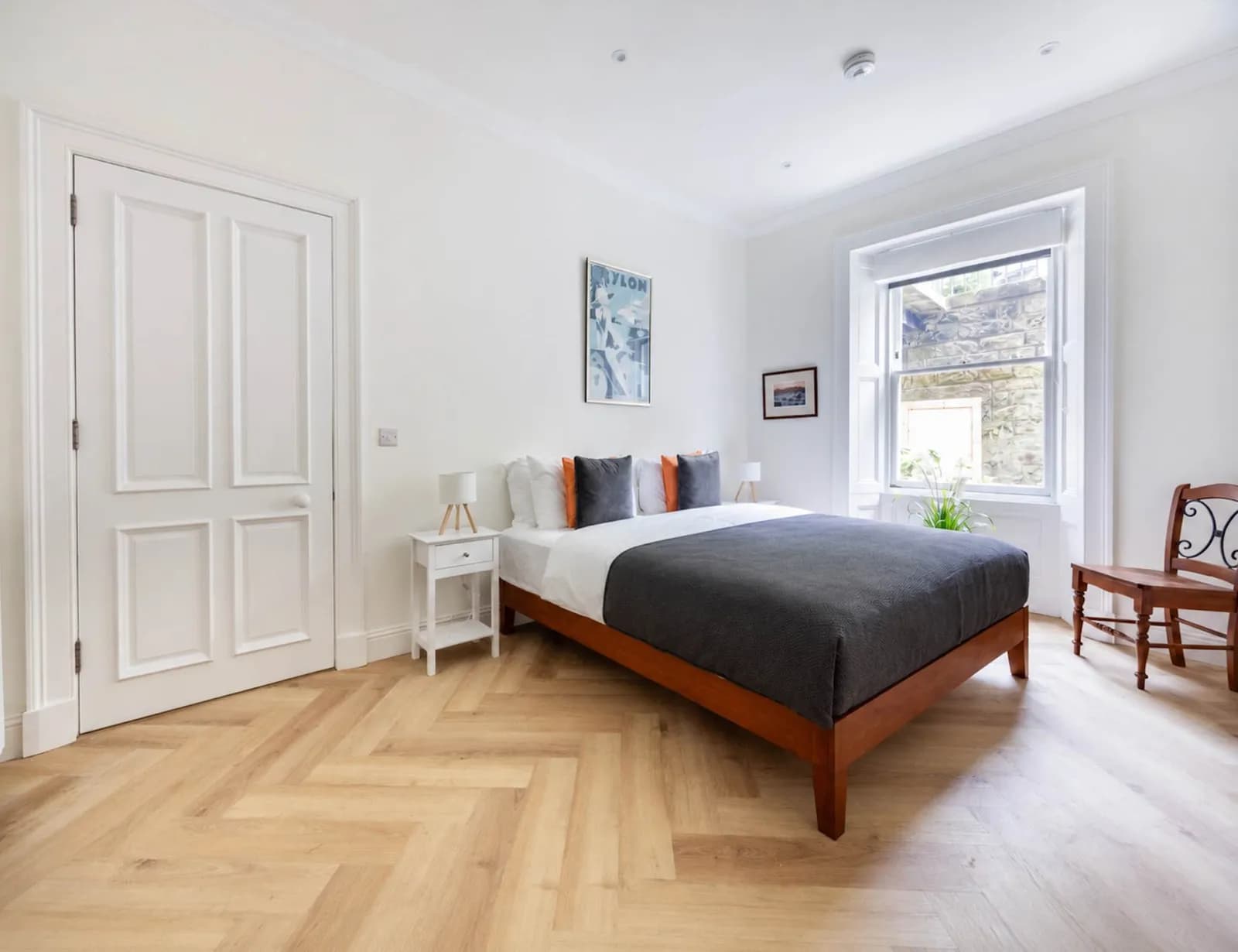 A clean, bright bedroom with white walls, a wooden bed with dark gray bedding, and a large window with white shutters.