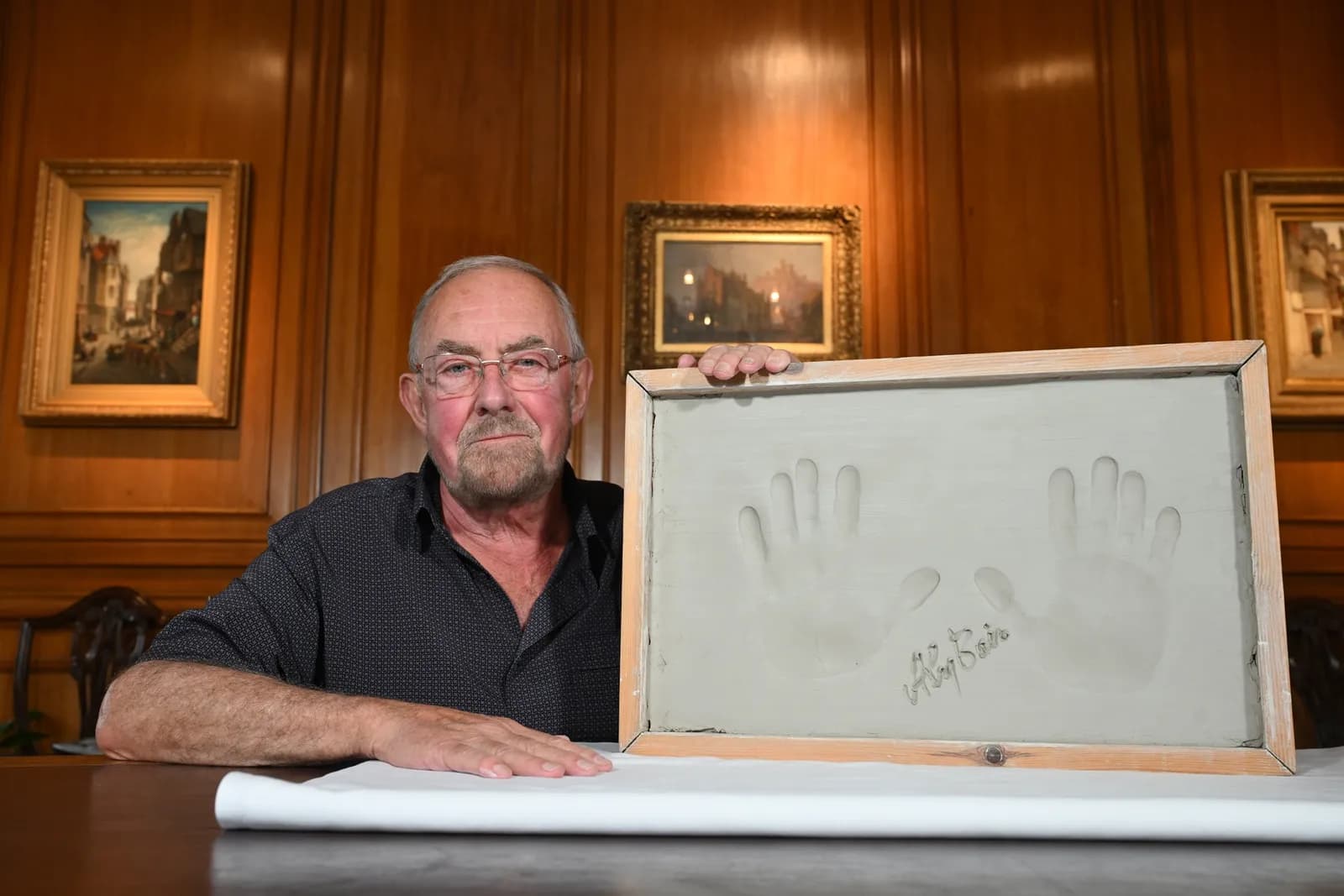 A man with a beard and glasses holds up a framed handprint with a signature on a table in a wood-paneled room.