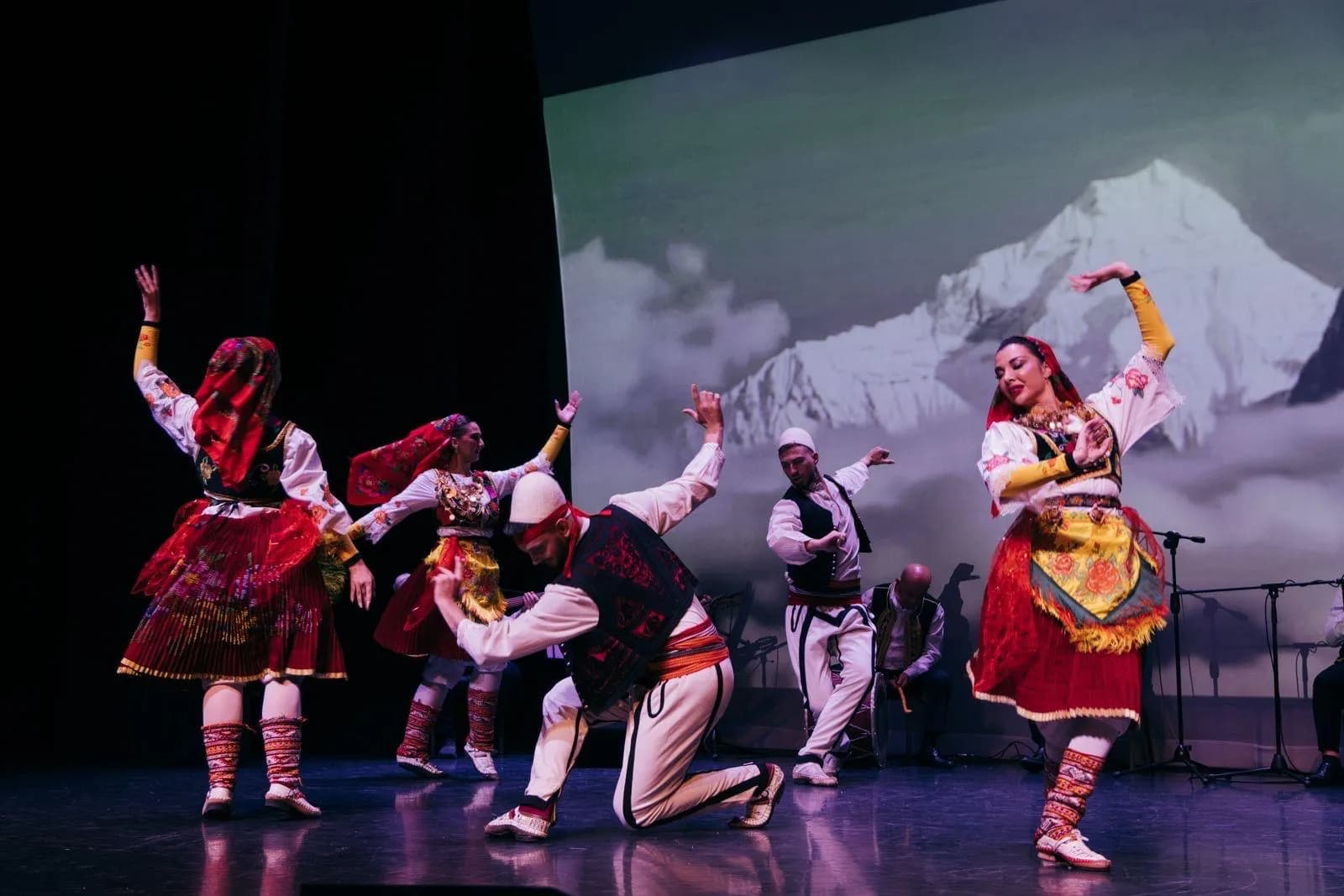 A group of dancers in traditional costumes performs on stage in front of a backdrop depicting snowy mountains and clouds.