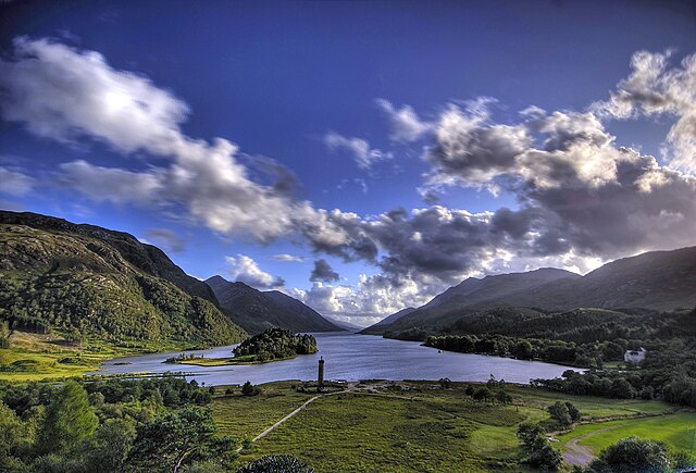 Glenfinnan Monument standing proudly beside the misty waters of Loch Shiel, framed by verdant hills under a cloudy sky.
